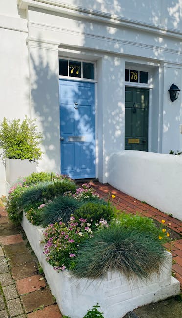 A daytime view of a charming front garden adjacent to a white building with two doors painted in pastel blue and dark green, each featuring a small window above. The garden bed, edged with white concr