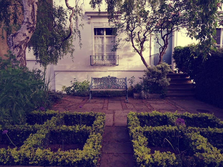 A daytime view of a charming front garden adjacent to a white building with two doors painted in pastel blue and dark green, each featuring a small window above. The garden bed, edged with white concr